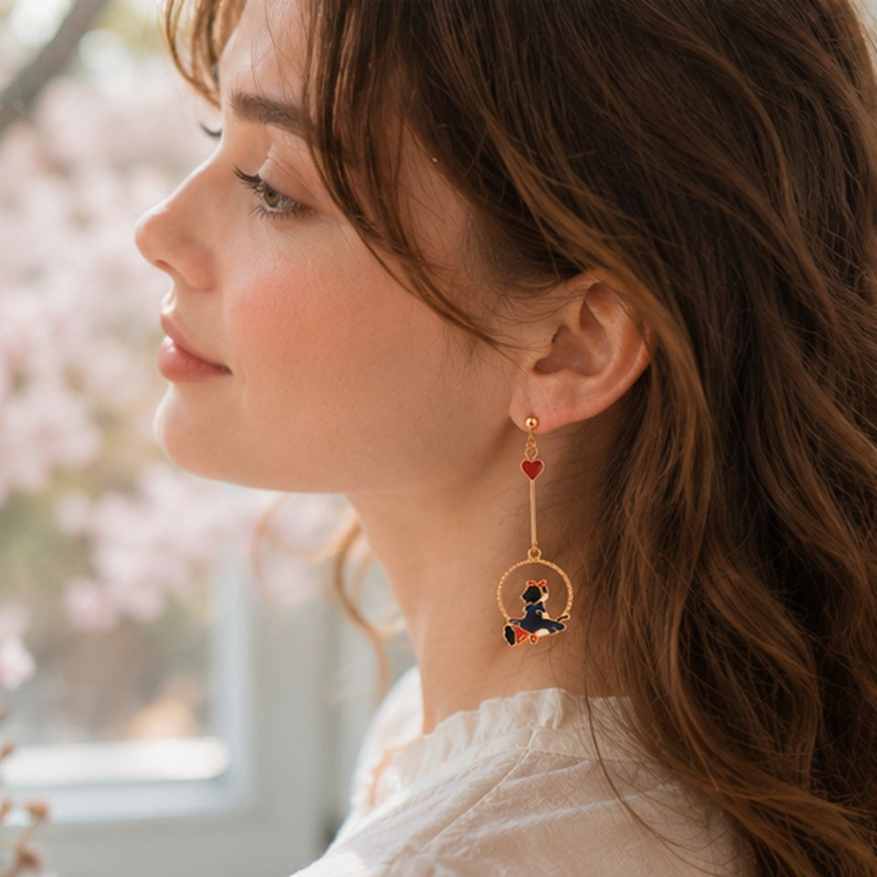 une paire de boucles d'oreilles asymétriques inspirées de Kiki la petite sorcière. D'un côté il y a Jiji le chat noir et de l'autre, Kiki sur son balai.