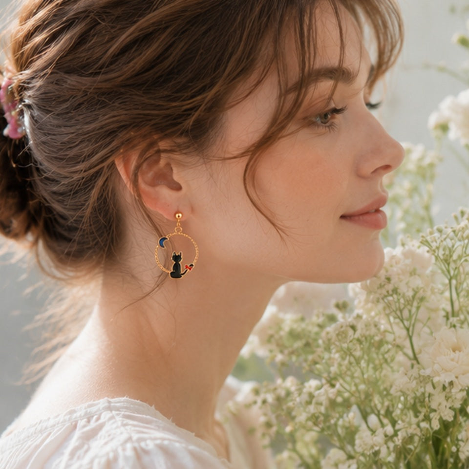 une paire de boucles d'oreilles asymétriques inspirées de Kiki la petite sorcière. D'un côté il y a Jiji le chat noir et de l'autre, Kiki sur son balai.