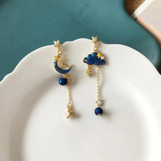 une paire de boucles d'oreilles asymétriques composée d'un côté d'un nuage et d'une perle bleue, et de l'autre, d'une lune et d'un strass en forme d'étoile