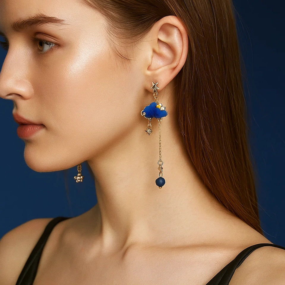 une paire de boucles d'oreilles asymétriques composée d'un côté d'un nuage et d'une perle bleue, et de l'autre, d'une lune et d'un strass en forme d'étoile
