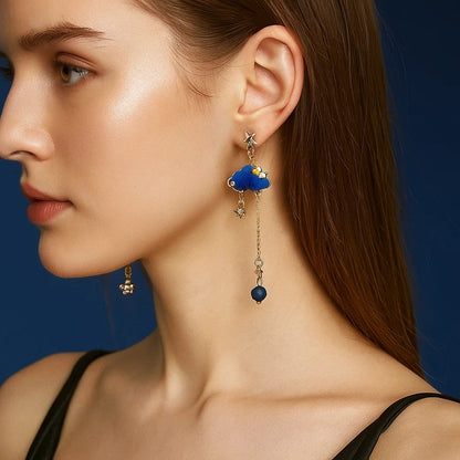 une paire de boucles d'oreilles asymétriques composée d'un côté d'un nuage et d'une perle bleue, et de l'autre, d'une lune et d'un strass en forme d'étoile