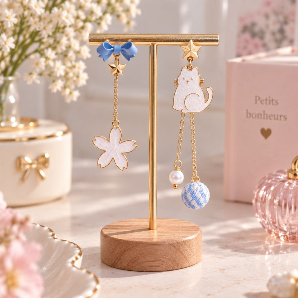Boucles d’oreilles asymétriques kawaii avec chat blanc, nœud bleu, fleur blanche et chaînes dorées, présentées sur un support à bijoux élégant.