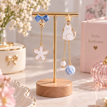 Boucles d’oreilles asymétriques kawaii avec chat blanc, nœud bleu, fleur blanche et chaînes dorées, présentées sur un support à bijoux élégant.