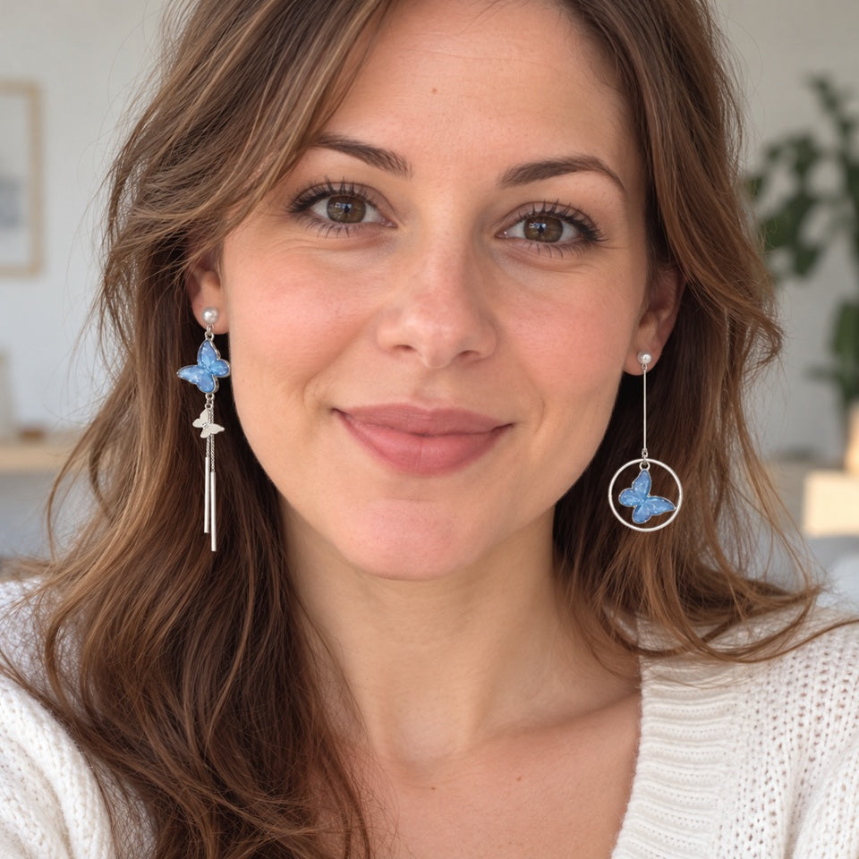 Boucles d’oreilles asymétriques argentées avec papillons bleus et perles blanches, portées par une femme, avec anneau et fines chaînettes pendantes.