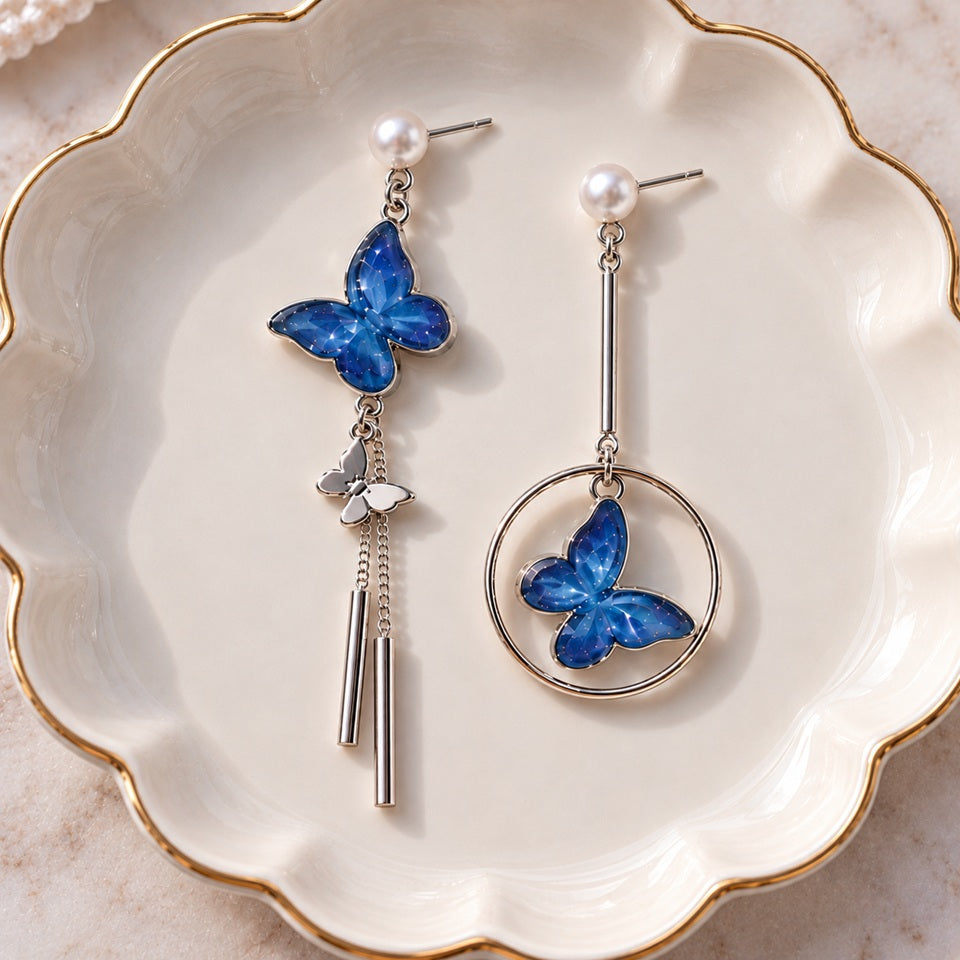 Boucles d’oreilles asymétriques argentées avec papillons bleus et perles blanches, composées d’un anneau et de fines chaînettes, présentées dans une coupelle élégante.