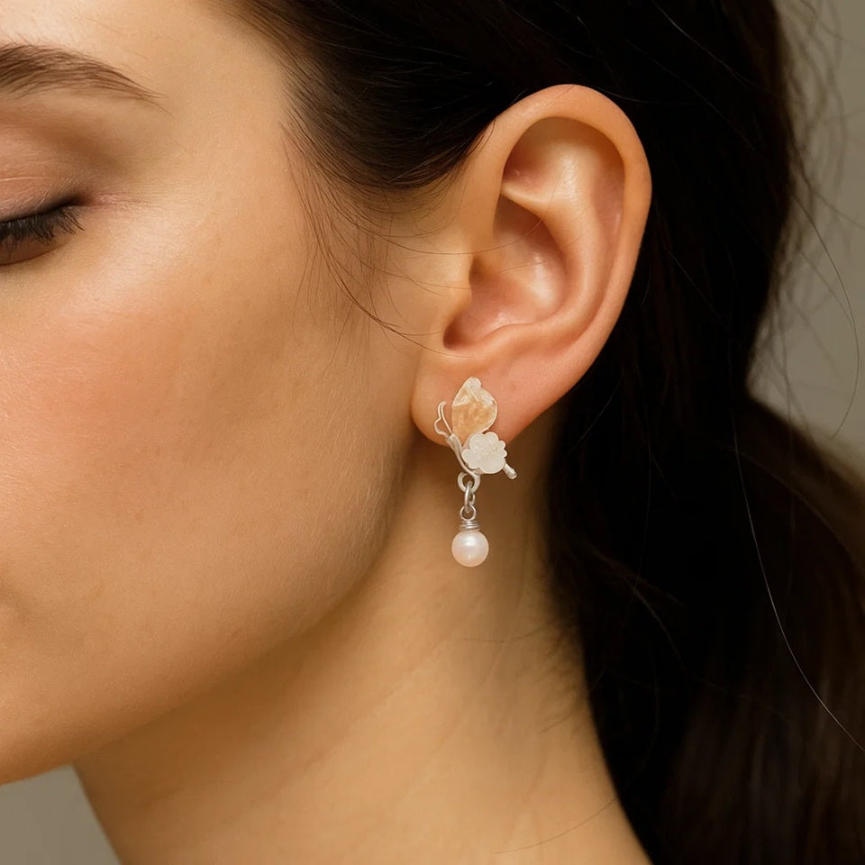 une paire de boucles d'oreilles florale et printanière représentant des papillons d'un rose pâle sur leurs ailes avec une jolie fleur blanche. Une perle blanche suspendu à leurs ailes vient compléter l'ensemble