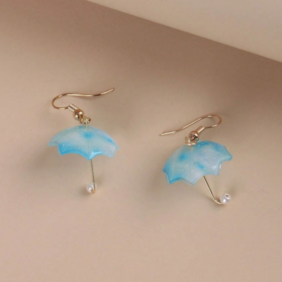 une paire de boucles d'oreilles en résine en forme de parapluie bleu avec un effet translucide. Au bout du manche, se trouve une perle blanche.