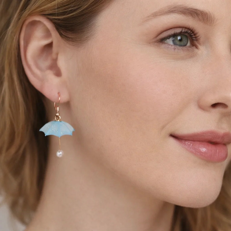 une paire de boucles d'oreilles en résine en forme de parapluie bleu avec un effet translucide. Au bout du manche, se trouve une perle blanche.