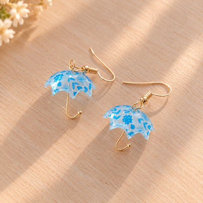 Paire de boucles d’oreilles fantaisie kawaii en forme de petits parapluies transparents bleu clair, décorés de délicates fleurs bleues. Leurs attaches et manches légèrement dorés apportent une touche élégante et raffinée. Posés sur une jolie table en bois clair, sous une douce lumière naturelle, ces bijoux pendants originaux au style romantique et floral sont parfaits pour compléter une tenue printanière avec une touche de poésie et de fantaisie.