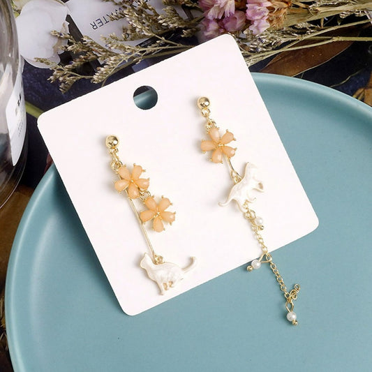 des boucles d'oreilles asymétriques et dorées. Un chat au ton nacré est posé tranquillement parmi des fleurs de sakura roses. L'ensemble est élégant et raffiné.