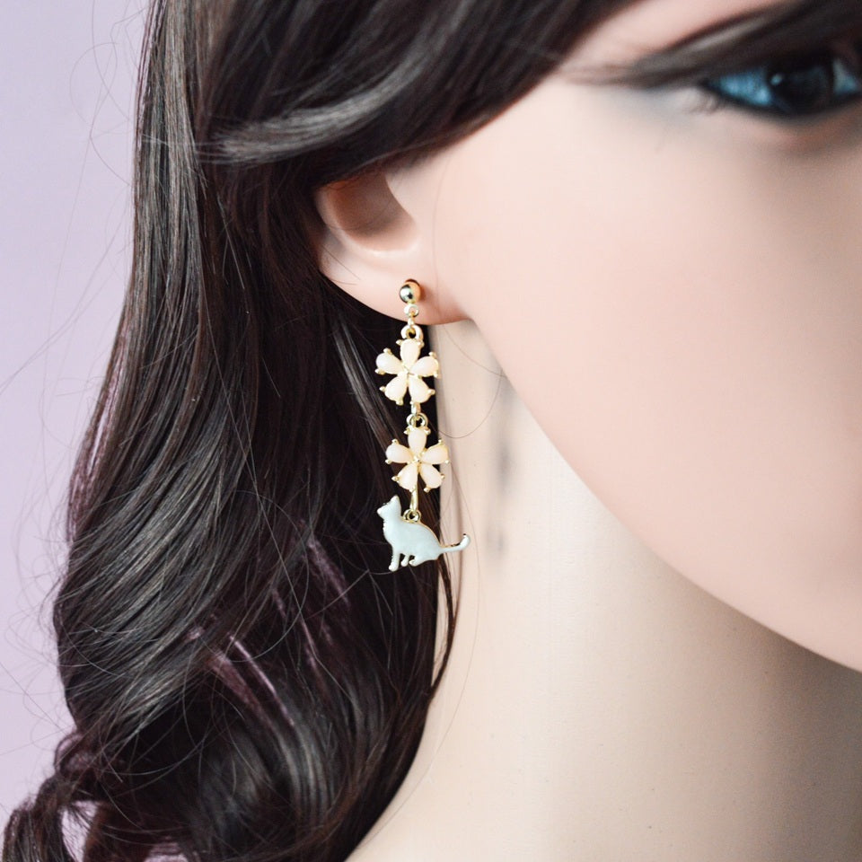 des boucles d'oreilles asymétriques et dorées. Un chat au ton nacré est posé tranquillement parmi des fleurs de sakura roses. L'ensemble est élégant et raffiné.