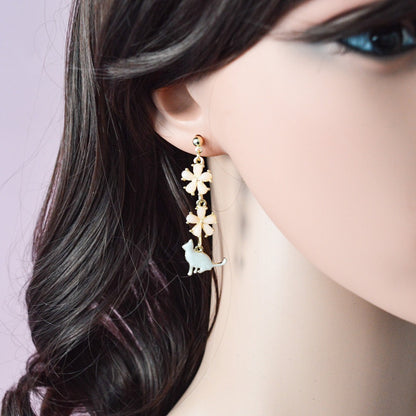 des boucles d'oreilles asymétriques et dorées. Un chat au ton nacré est posé tranquillement parmi des fleurs de sakura roses. L'ensemble est élégant et raffiné.