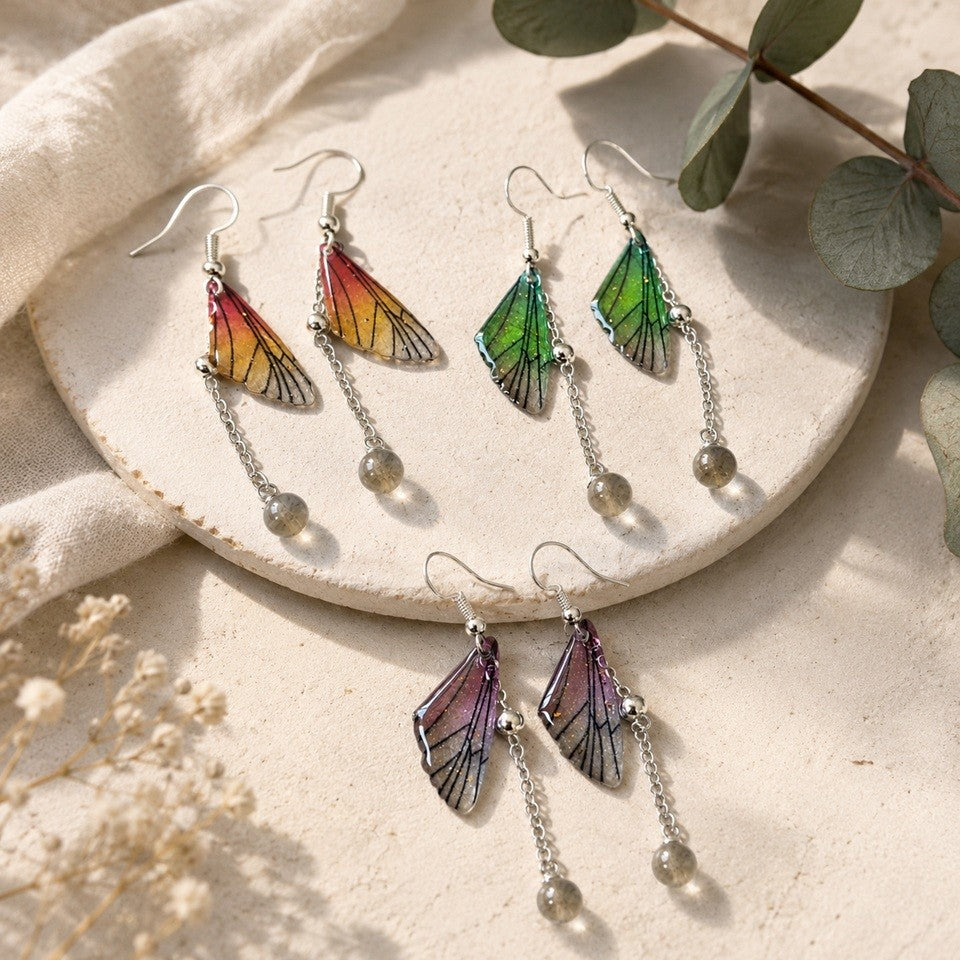 Mockup carré présentant trois paires de boucles d'oreilles artisanales inspirées d'ailes de papillon, déclinées en orange, vert et violet. Les boucles, montées sur crochets argentés avec une fine chaîne terminée par une perle translucide, sont disposées sur un plateau en céramique beige entouré d'un tissu naturel, de feuilles d'eucalyptus et de fleurs séchées. Une lumière douce met en valeur les couleurs délicates et les détails des ailes, créant une ambiance élégante et naturelle.