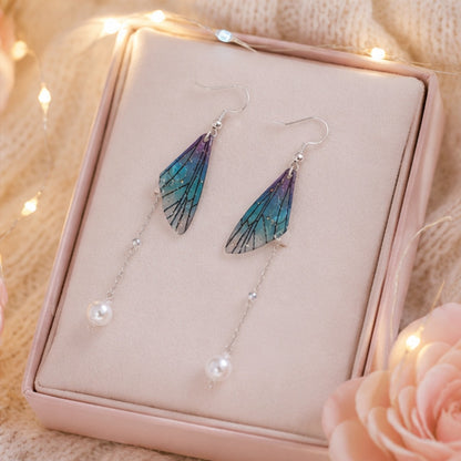 Des boucles d'oreilles pendantes en forme d'aile de papillon bleu-violet avec perle blanche, posées sur une boite à bijoux rose dans un décor romantique lumineux et doux.