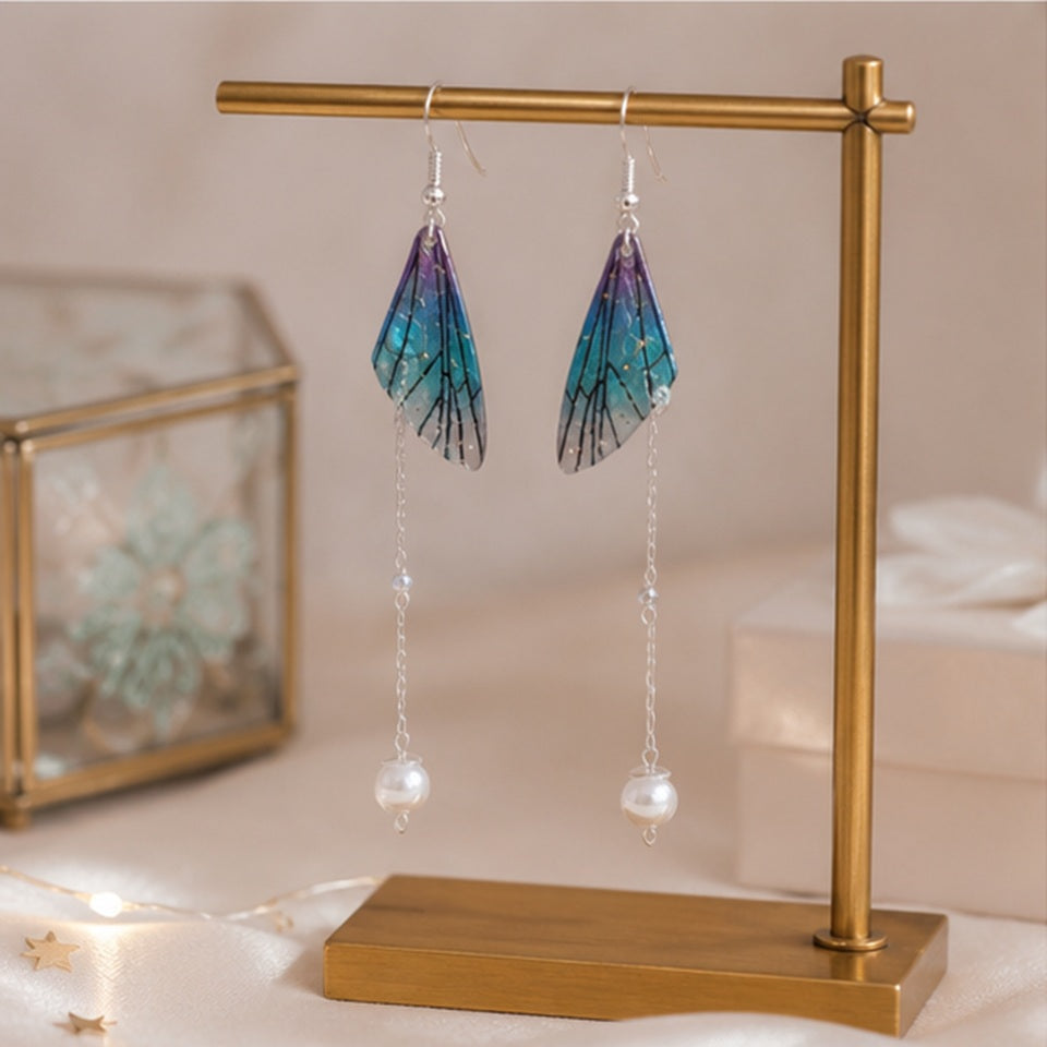 des boucles d'oreilles pendantes en forme d'aile de papillon bleu-violet avec perle blanche accrochées à un support de bijouterie doré, sur une coiffeuse féminine rose.