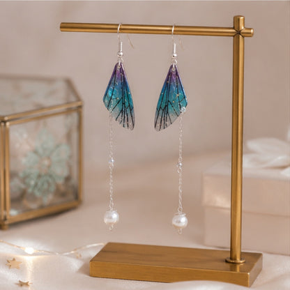 des boucles d'oreilles pendantes en forme d'aile de papillon bleu-violet avec perle blanche accrochées à un support de bijouterie doré, sur une coiffeuse féminine rose.