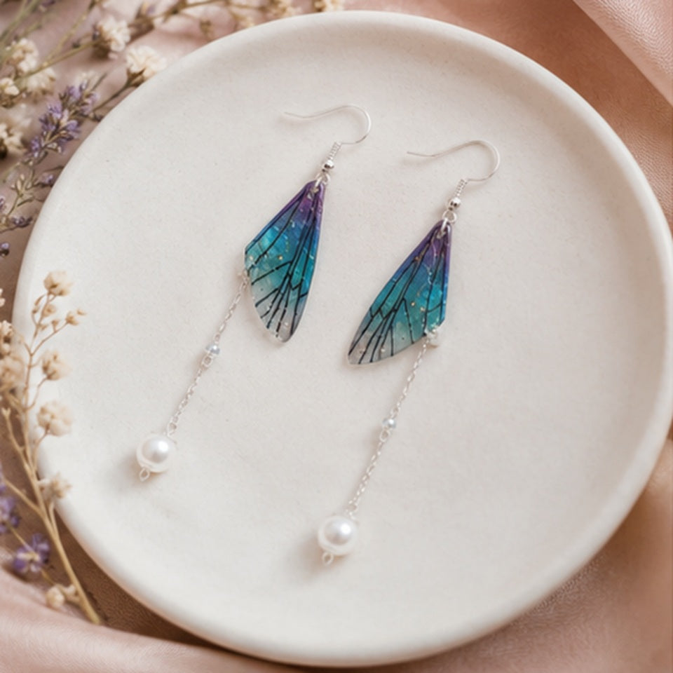 Des boucles d'oreilles pendantes en forme d'aile de papillon bleu violet avec perle blanche, posées dans une assiette ronde blanche sur une jolie nappe rose doré décorée de fleurs séchées.