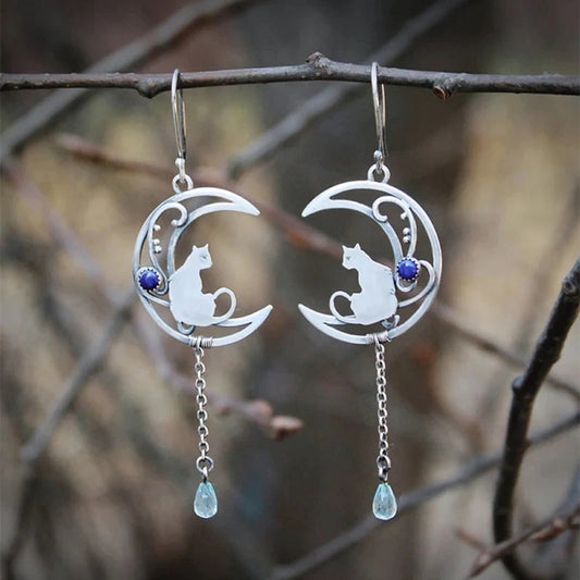 des boucles d'oreilles argentées en forme de lune ornée d'une perle bleue. Un chat repose sur la lune et une chainette y est suspendue. Une perle en forme de goutte bleue se trouve au bout.