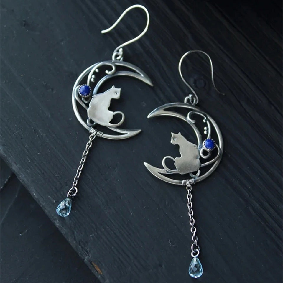 des boucles d'oreilles argentées en forme de lune ornée d'une perle bleue. Un chat repose sur la lune et une chainette y est suspendue. Une perle en forme de goutte bleue se trouve au bout.