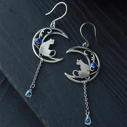des boucles d'oreilles argentées en forme de lune ornée d'une perle bleue. Un chat repose sur la lune et une chainette y est suspendue. Une perle en forme de goutte bleue se trouve au bout.