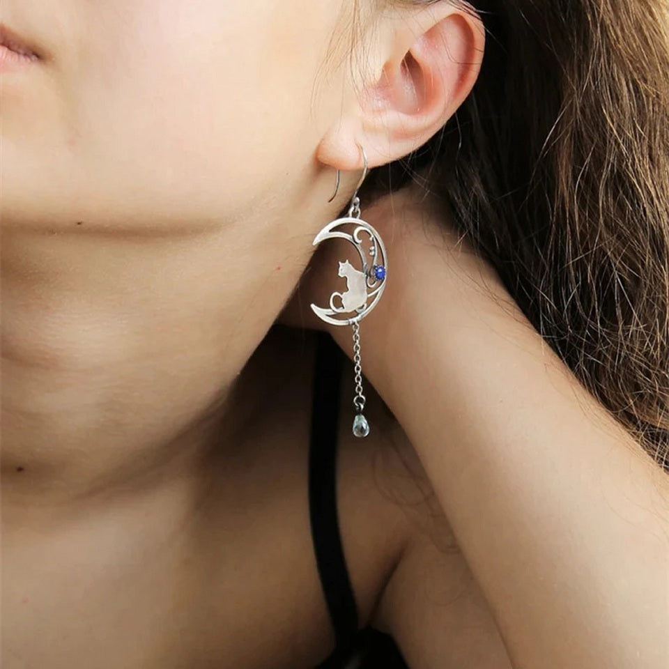 des boucles d'oreilles argentées en forme de lune ornée d'une perle bleue. Un chat repose sur la lune et une chainette y est suspendue. Une perle en forme de goutte bleue se trouve au bout.