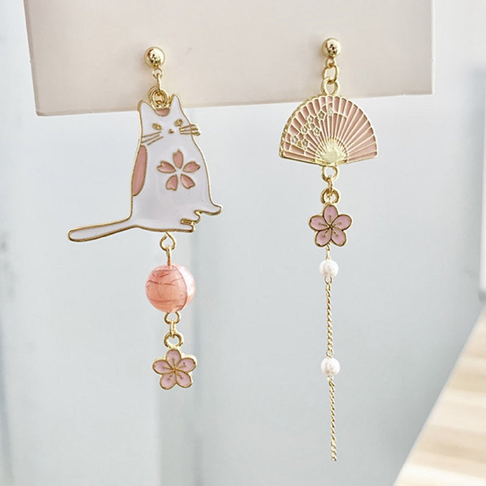 des boucles d'oreilles asymétriques avec un chat blanc et une perle rose d'un côté. De l'autre côté, il y  a un éventail avec une chainette ornée de perles blanches et d'une petite fleur de sakura.