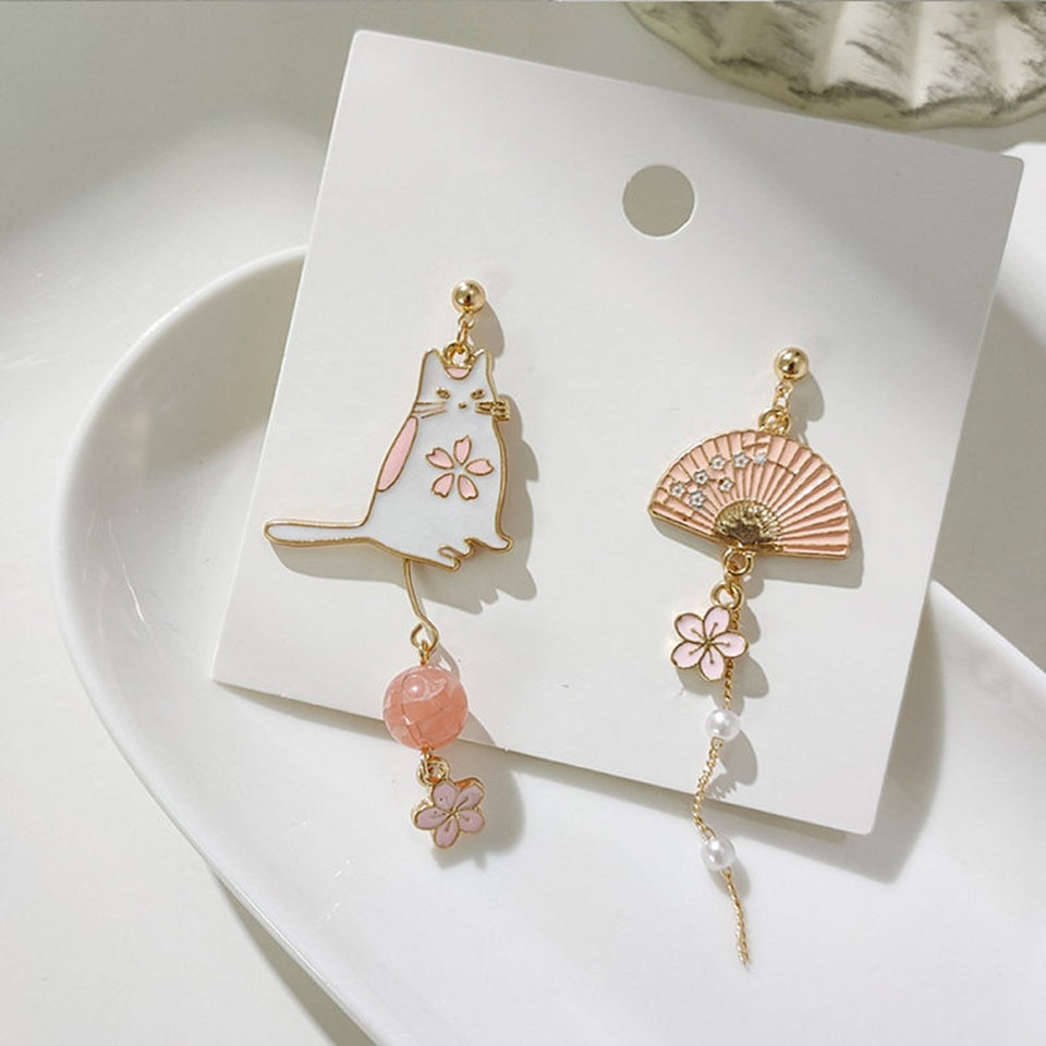 des boucles d'oreilles asymétriques avec un chat blanc et une perle rose d'un côté. De l'autre côté, il y  a un éventail avec une chainette ornée de perles blanches et d'une petite fleur de sakura.