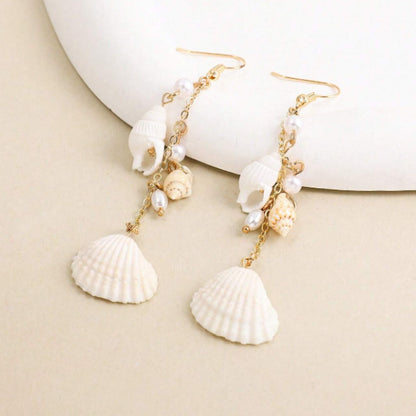 des boucles d'oreilles longues et dorées formées de divers coquillages blancs. L'ensemble évoque les vacances, la plage et l'été.