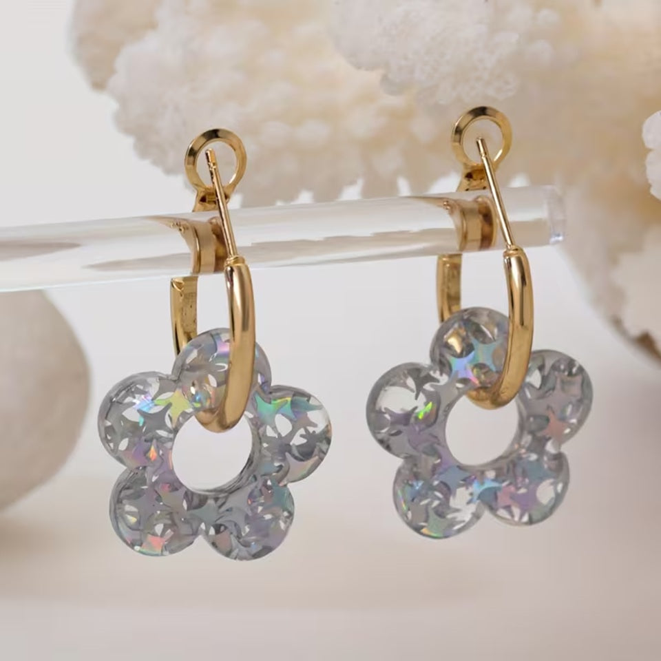 gros plan sur une paire de boucles d'oreilles créoles dorées dans lesquelles passent une joli fleur scintillante en acrylique. Elle a des paillettes argentés qui captent et reflètent la lumière pour créer de jolis effets lumineux. Style bohème chic assuré !