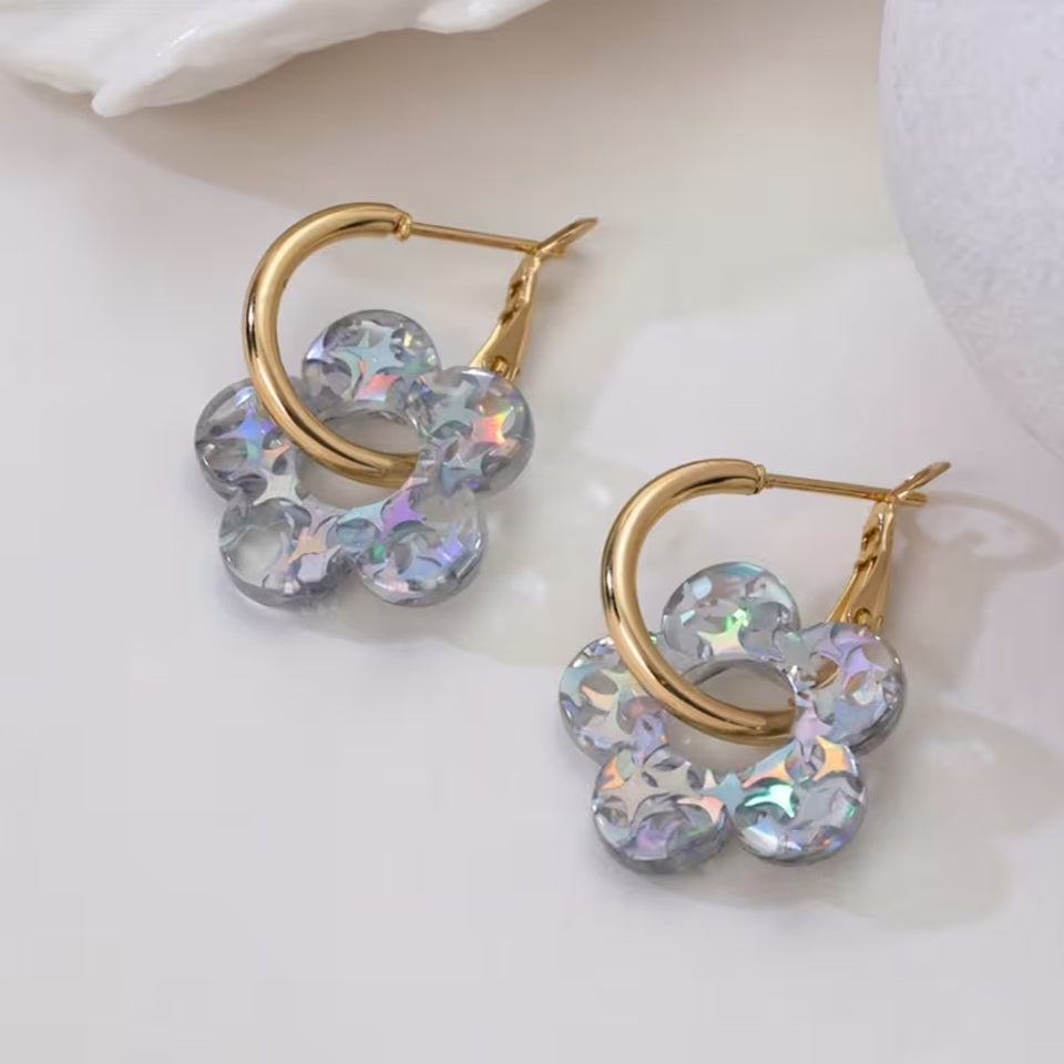gros plan sur une paire de boucles d'oreilles créoles dorées dans lesquelles passent une joli fleur scintillante en acrylique. Elle a des paillettes argentés qui captent et reflètent la lumière pour créer de jolis effets lumineux.