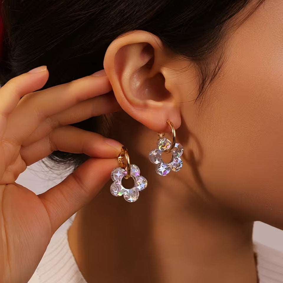 Une femme brune porte à son oreille une boucle d'oreille créole dorée dans laquelle passe une joli fleur scintillante en acrylique. Elle a des paillettes argentés qui captent et reflètent la lumière pour créer de jolis effets lumineux.