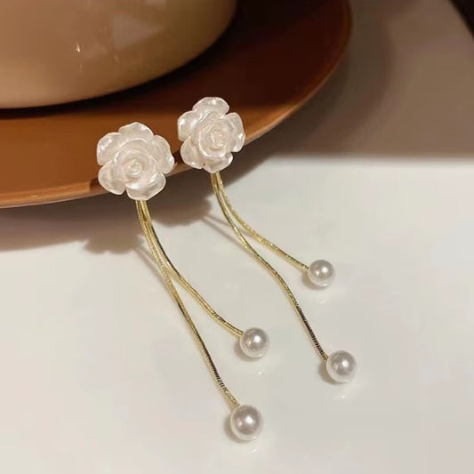 une paire de boucles d'oreilles en forme de roses blanches d'où pendent une délicate perle blanche. En blanc et doré, l'ensemble est harmonieux et délicat