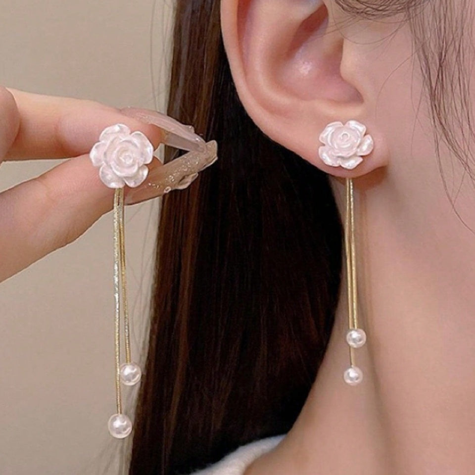 une paire de boucles d'oreilles en forme de roses blanches d'où pendent une délicate perle blanche. En blanc et doré, l'ensemble est harmonieux et délicat