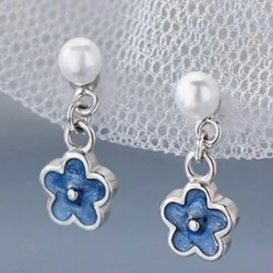 une paire de boucles d'oreilles composées d'une perle blanche et d'une petite fleur bleue, l'ensemble est romantique et bohème