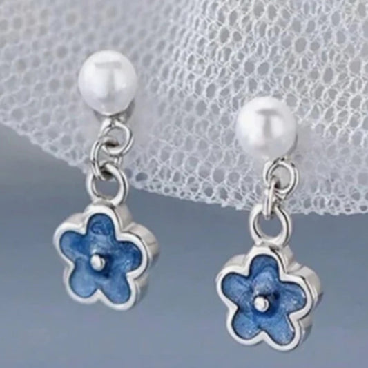 une paire de boucles d'oreilles composées d'une perle blanche et d'une petite fleur bleue, l'ensemble est romantique et bohème