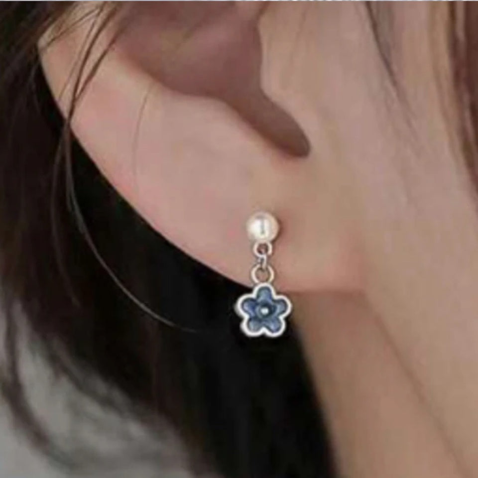 une paire de boucles d'oreilles composées d'une perle blanche et d'une petite fleur bleue, l'ensemble est romantique et bohème