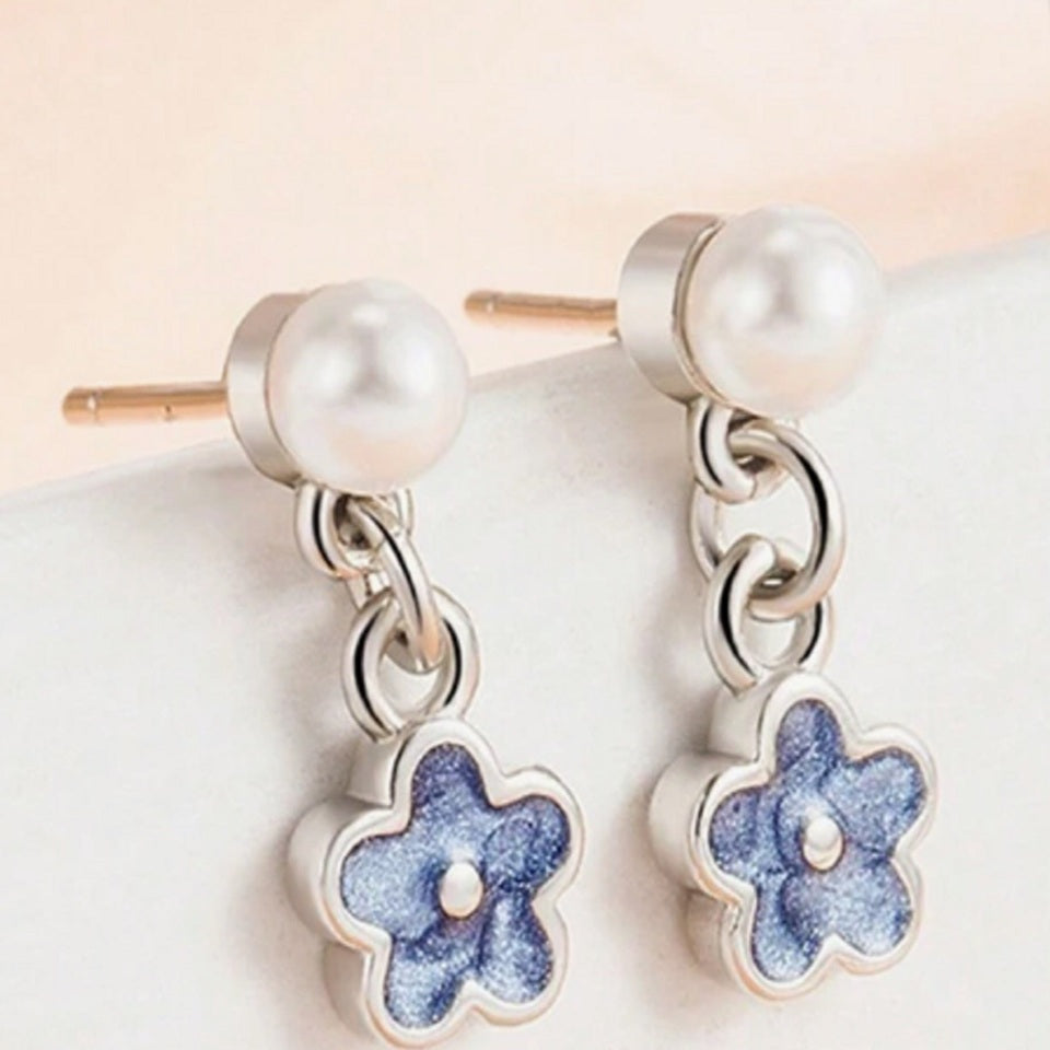une paire de boucles d'oreilles composées d'une perle blanche et d'une petite fleur bleue, l'ensemble est romantique et bohème