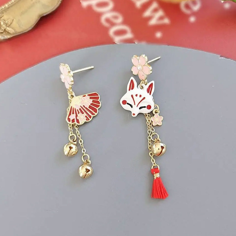 une paire de boucles d'oreilles asymétriques inspirées du folklore japonais. d'un côté un kitsune  avec un petit pompon rouge, de l'autre, un éventail. L'ensemble des couleurs rouge et blanc forment une harmonie parfaite. Des petites clochettes viennent compléter le tout.