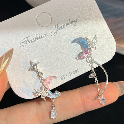 des boucles d'oreilles asymétriques en forme de lune bicolore bleue et rose sur laquelle est posé un papillon argenté. Une chainette y est suspendue au bout de laquelle pend un strass enforme de goutte.
