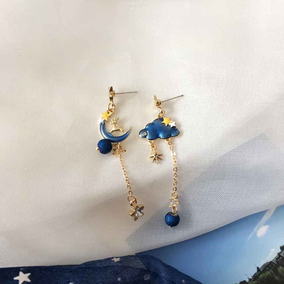 une paire de boucles d'oreilles asymétriques composée d'un côté d'un nuage et d'une perle bleue, et de l'autre, d'une lune et d'un strass en forme d'étoile