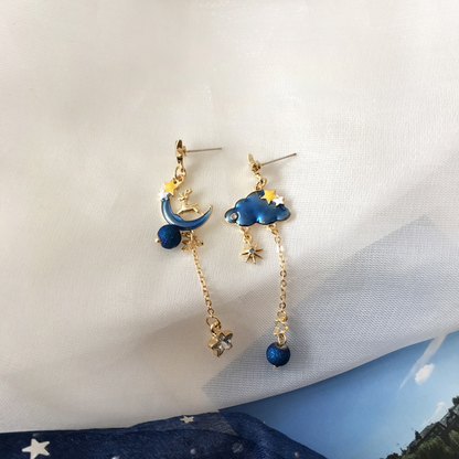 une paire de boucles d'oreilles asymétriques composée d'un côté d'un nuage et d'une perle bleue, et de l'autre, d'une lune et d'un strass en forme d'étoile