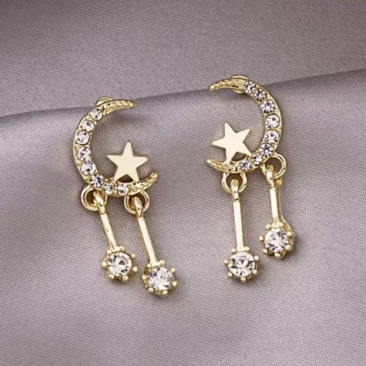 une paire de boucles d'oreilles en forme de lune toute en strass dans laquelle repose une étoile dorée. Deux strass sont suspendus à la lune et apportent encore plus de brillance.