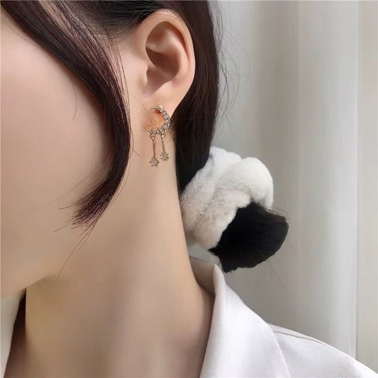 une paire de boucles d'oreilles en forme de lune toute en strass dans laquelle repose une étoile dorée. Deux strass sont suspendus à la lune et apportent encore plus de brillance.