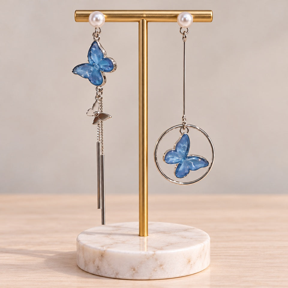 Boucles d’oreilles asymétriques argentées avec papillons bleus, chaînes fines et anneau, présentées sur un élégant support à bijoux doré.