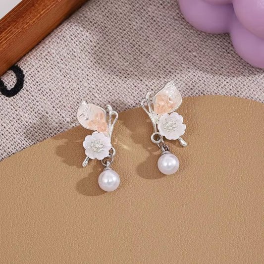 une paire de boucles d'oreilles florale et printanière représentant des papillons d'un rose pâle sur leurs ailes avec une jolie fleur blanche. Une perle blanche suspendu à leurs ailes vient compléter l'ensemble