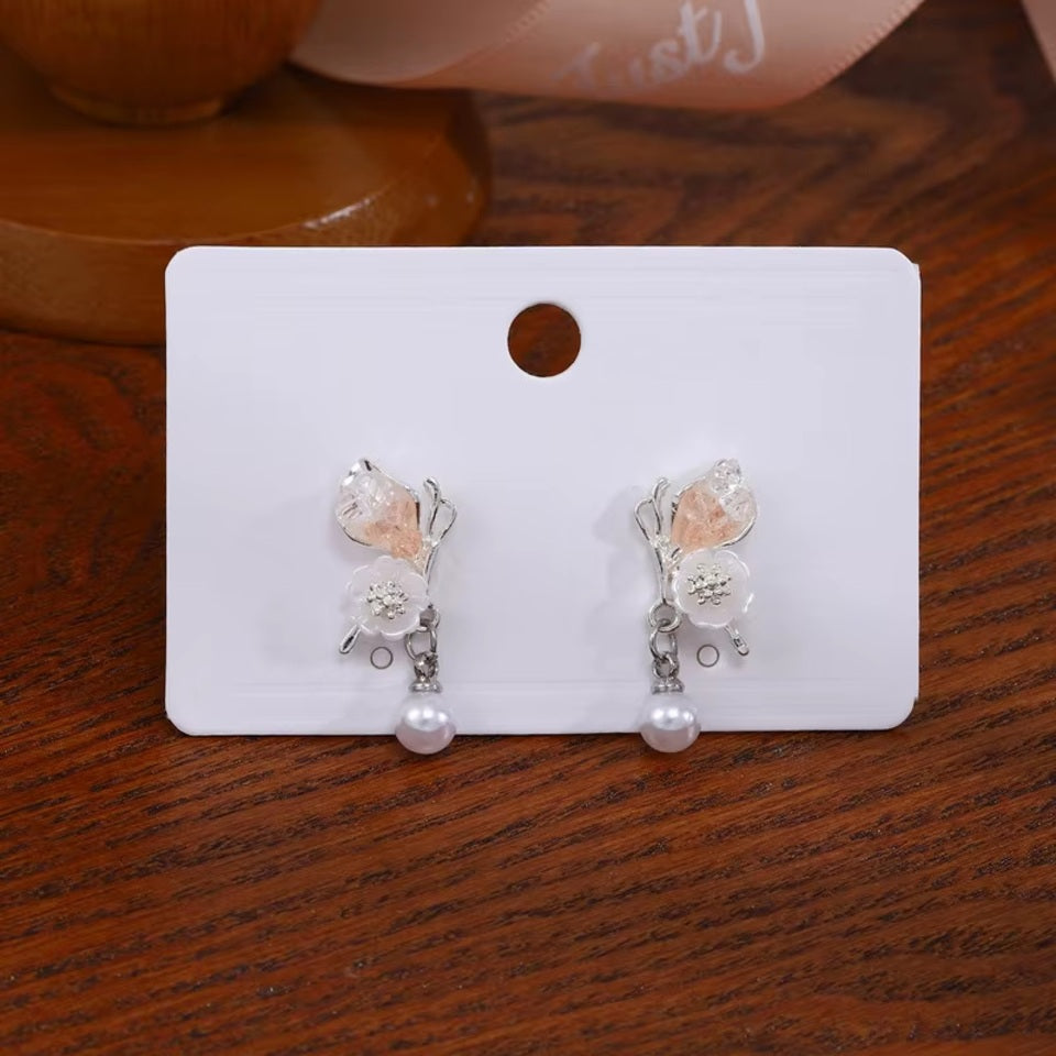 une paire de boucles d'oreilles florale et printanière représentant des papillons d'un rose pâle sur leurs ailes avec une jolie fleur blanche. Une perle blanche suspendu à leurs ailes vient compléter l'ensemble