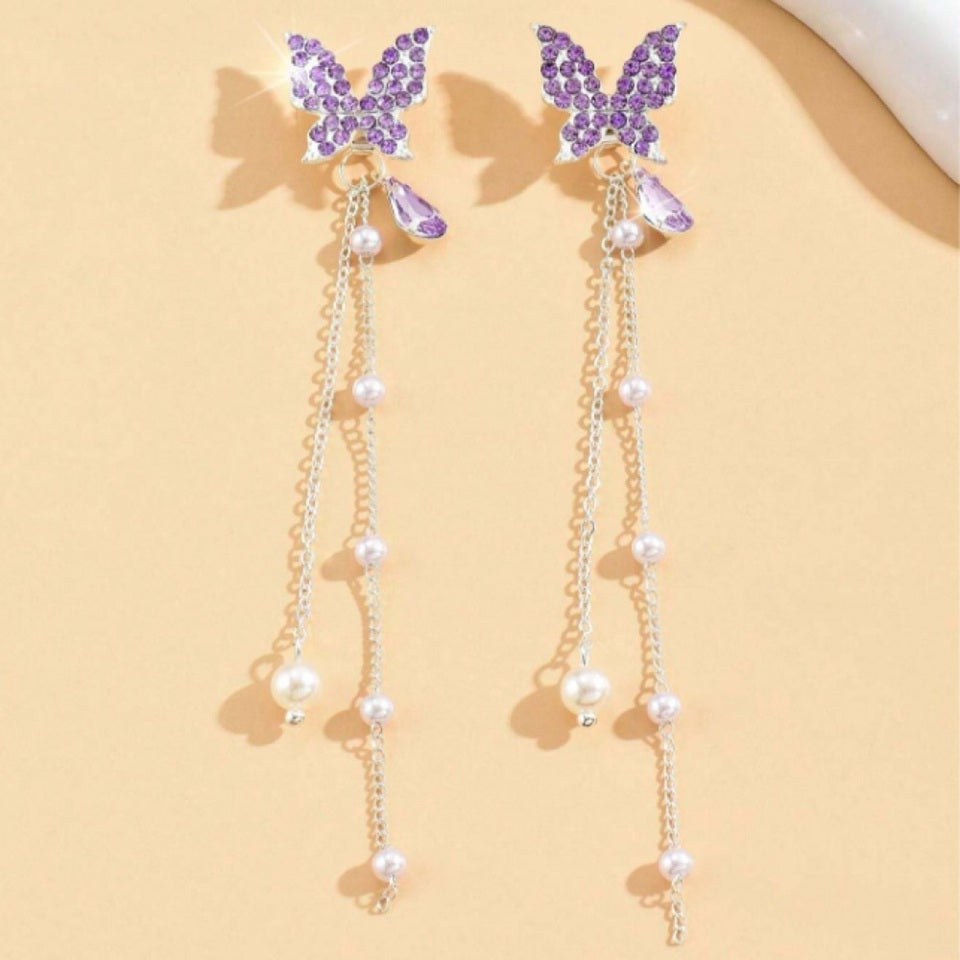 des boucles d'oreilles en forme de papillons violets tout en strass. Des chainettes de perles blanches y sont suspendus