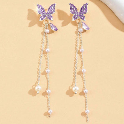 des boucles d'oreilles en forme de papillons violets tout en strass. Des chainettes de perles blanches y sont suspendus
