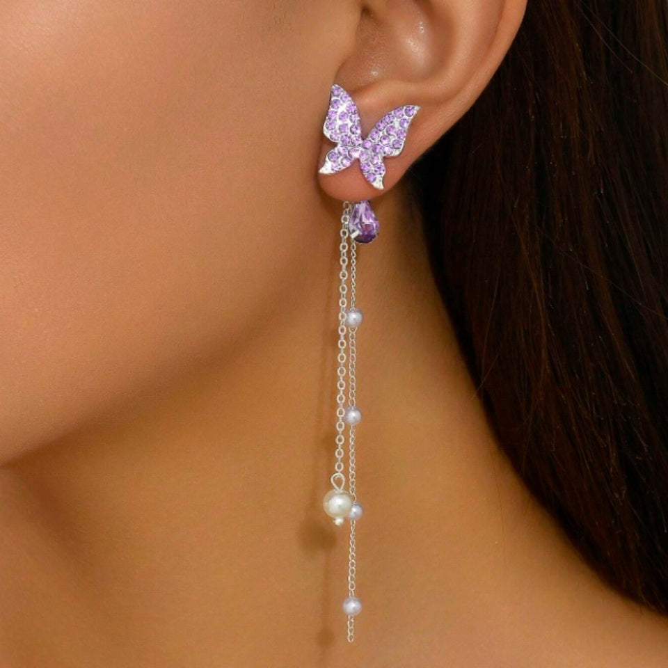 des boucles d'oreilles en forme de papillons violets tout en strass. Des chainettes de perles blanches y sont suspendus