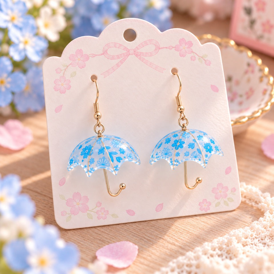 Paire de boucles d’oreilles fantaisie en forme de petits parapluies transparents, décorés de délicates fleurs bleues et de motifs floraux. Leurs fines attaches et manches légèrement dorés apportent une touche élégante et raffinée. Présentées sur un joli support rose pastel orné de fleurs de sakura, dans un décor romantique aux tons doux, ces boucles d’oreilles kawaii originales sont idéales pour compléter une tenue printanière, féminine et poétique.